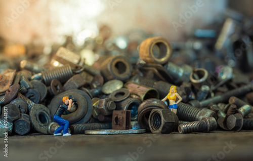 Miniature male photographer standing to take photo a female model on pile of old screws nuts and bolts