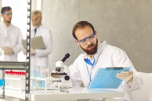 Portrait of man scientist in eyeglasses and lab coat sitting at desk looking at clipboard with test result analyzing sample using microscope. Research group working at laboratory on background
