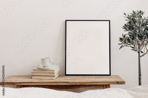 Vertical black picture frame mockup on vintage bench, table. Cup of coffee on pile of books. Potted olive tree. White wall background. Scandinavian interior, neutral color palette. Selective focus.