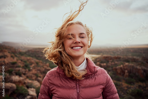 Smiling young confident woman with closed eyes feeling fresh wind against face standing on mountain hill photo