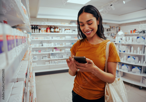 Fotografie Young woman smiling while using smartphone standing against shelf in pharmacy