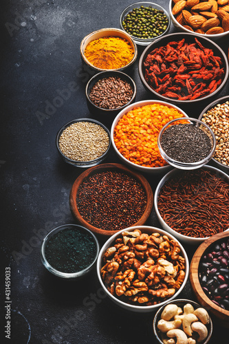 Selection of superfoods, legumes, cereals, nuts, seeds in bowls on black background. Superfood as chia, spirulina, beans, goji berries, quinoa, turmeric and other. Copy space, top view