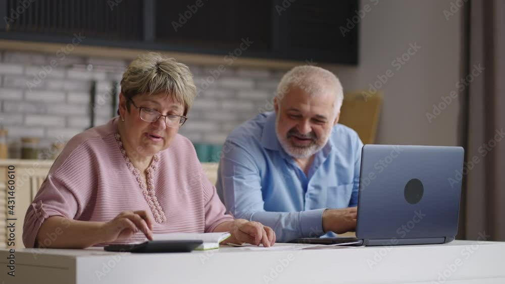 old man and woman are paying utility bills online, sitting at kitchen together, wife is calculating and man is using online payment service old man and woman are paying utility bills online, sitting at kitchen together, wife is calculating and man is using online payment service