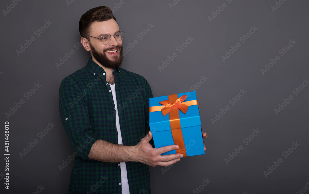 Happy young handsome bearded man in casual wear in good mood with big gift box in his hands having fun and posing over gray background Happy young handsome bearded man in casual wear in good mood with big gift box in his hands having fun and posing over gray background