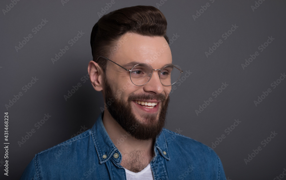Fototapeta premium Confident smart modern bearded man in casual denim clothes and eyewear is posing over gray background in the studio