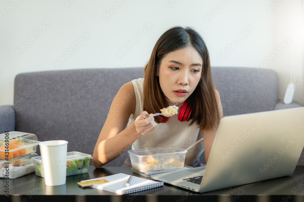 Portrait of young Asian woman freelancer working with laptop at her home and eating Asian food with takeaway. Work at home during self-isolation quarantine and New normal concept. Portrait of young Asian woman freelancer working with laptop at her home and eating Asian food with takeaway. Work at home during self-isolation quarantine and New normal concept.