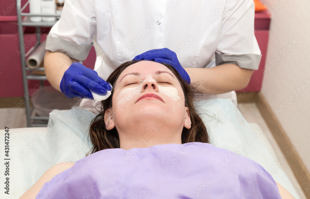 Application of cosmetology mask on the face of a young woman at spa salon. Cosmetologist hand in blue protective glove touching patient face with cotton pad Application of cosmetology mask on the face of a young woman at spa salon. Cosmetologist hand in blue protective glove touching patient face with cotton pad