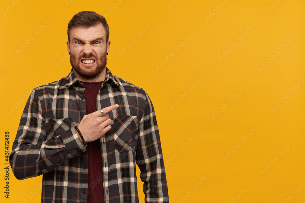 Angry irritated young man in plaid shirt with beard standing and pointing by finger away to the side over yellow background Angry irritated young man in plaid shirt with beard standing and pointing by finger away to the side over yellow background