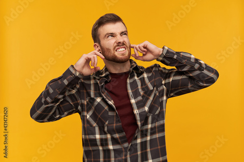 Annoyed angry young man in checkered shirt with beard covered ears by fingers and feels irritated beacause of noise over yellow background photo