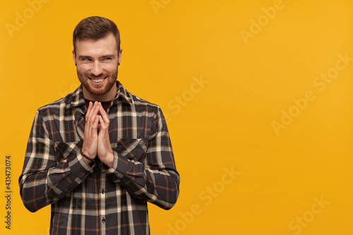 Mischievous arrogant young man in plaid shirt with beard rubbing hands and scheming an evil plan over yellow background Looking away to the side photo