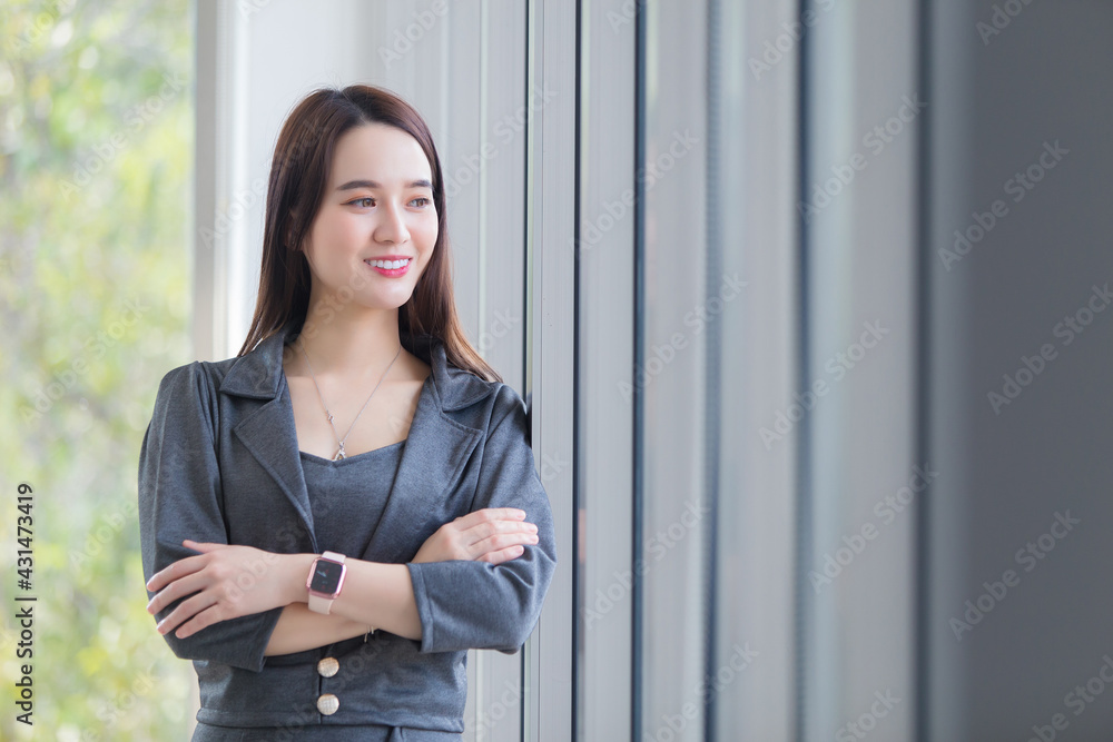 Asian business woman who has long hair wears grey dress and arms cross  while she is looking out from window and smiling happily.
