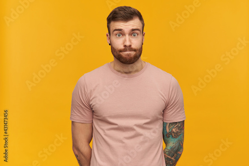 Shy embarrassed tattooed young bearded man in pink tshirt keeps hands behind himself and looks timid over yellow background