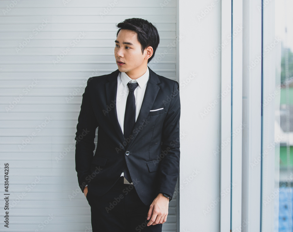 Young asian businessman formal wear in black. Standing right hand in pant pocket looking beside in white office room. Looks confident smart leadership relaxed style. Have space for text, marketing. Young asian businessman formal wear in black. Standing right hand in pant pocket looking beside in white office room. Looks confident smart leadership relaxed style. Have space for text, marketing.