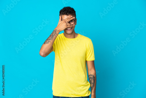 Young Brazilian man isolated on blue background covering eyes by hands and smiling