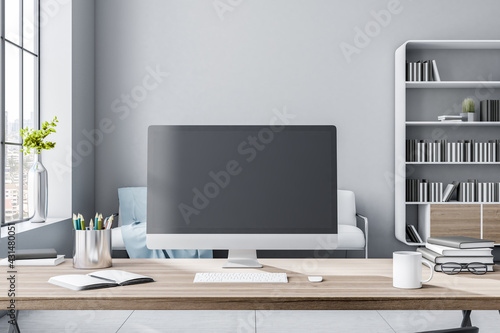 Black computer monitor with copyspace on wooden table in sunny room with big window and bookcase on background. 3D rendering, mock up