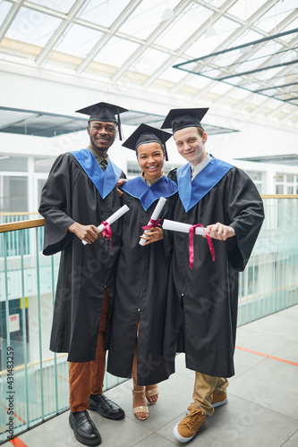 Vertical full length view at diverse group of college graduates holding diploma certificates and smiling at camera indoors