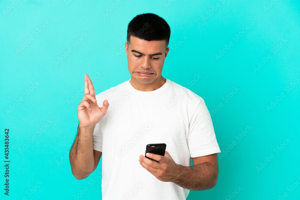 Young handsome man over isolated blue background using mobile phone with fingers crossing Young handsome man over isolated blue background using mobile phone with fingers crossing