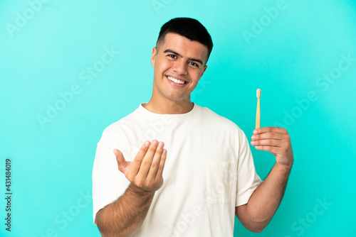 Young handsome man brushing teeth over isolated blue background inviting to come with hand. Happy that you came