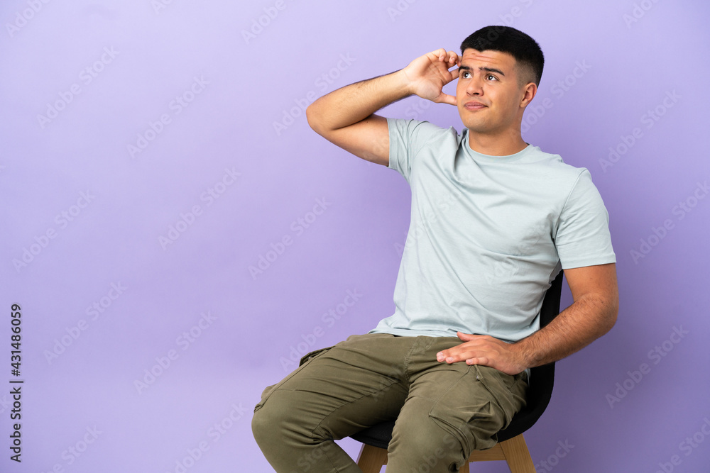 Young man sitting on a chair over isolated background having doubts and with confuse face expression Young man sitting on a chair over isolated background having doubts and with confuse face expression