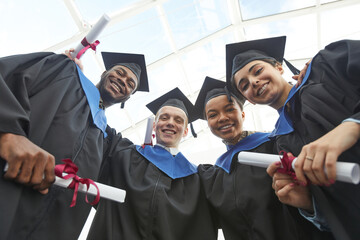 Low angle view at diverse group of happy young people wearing graduation gowns and holding diplomas while looking down at camera