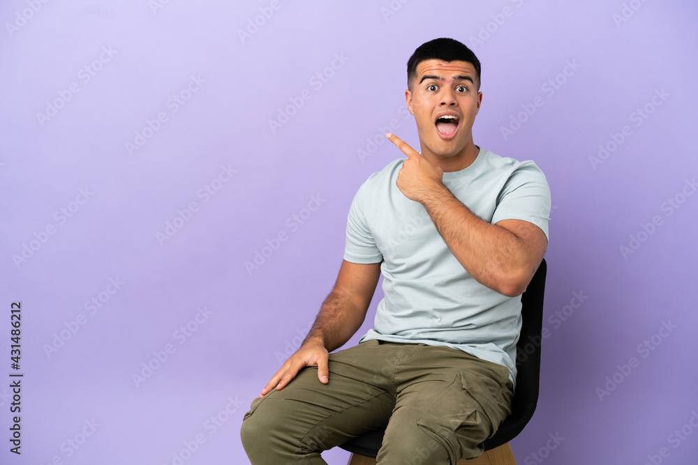Young man sitting on a chair over isolated background surprised and pointing side Young man sitting on a chair over isolated background surprised and pointing side