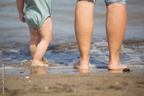 Baby legs and feet with mum legs on the shore of a beach while getting wet in cold water on a summer family holidays scene