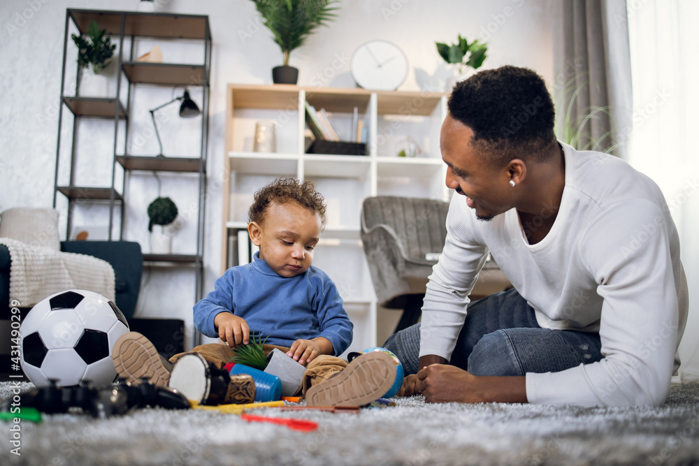 Young afro american father taking care of his cute son while staying together at home. Happy man and baby boy in casual clothes sitting on carpet and playing with toys.