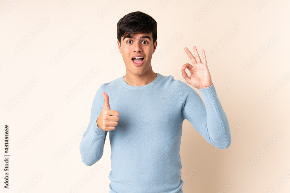 Handsome man over isolated beige background showing ok sign and thumb up gesture Handsome man over isolated beige background showing ok sign and thumb up gesture