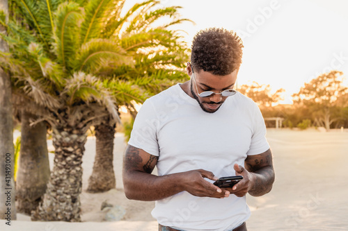 Portrait of a handsome black and young american man wearing sunglasses and using a smartphone during sunset at the beach with palms in the background