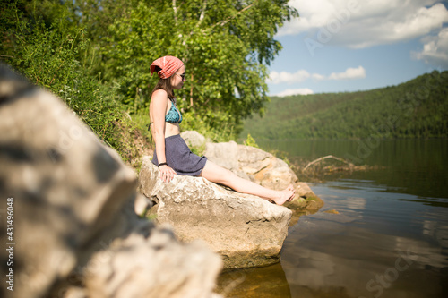 A young white beautiful staunch girl in a swimsuit and a short skirt sits on a rock on the river bank and enjoys the sun. She tans and smiles, she has a scarf on her head
