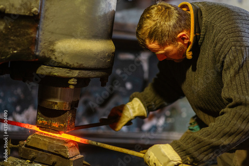 A blacksmith forges an iron rod with a mechanical hammer. Work in the forge