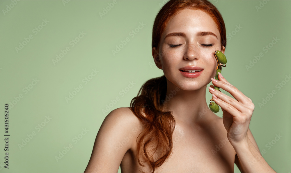 Obraz na płótnie Beauty spa. Redhead woman massage facial skin with natural jade roller. Girl pleasure from using lifting tool on her face, massaging muscles to reduce tension, rubbing in c-serum skincare treatment w salonie