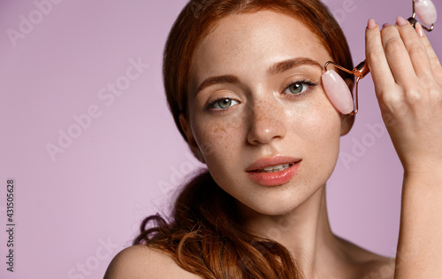 Skincare spa. Close up redhead woman uses natural quarz roller under eye, reduce puffiness and dark circles. Female self massage facial skin, after cleansing and nourishing face, pink background
