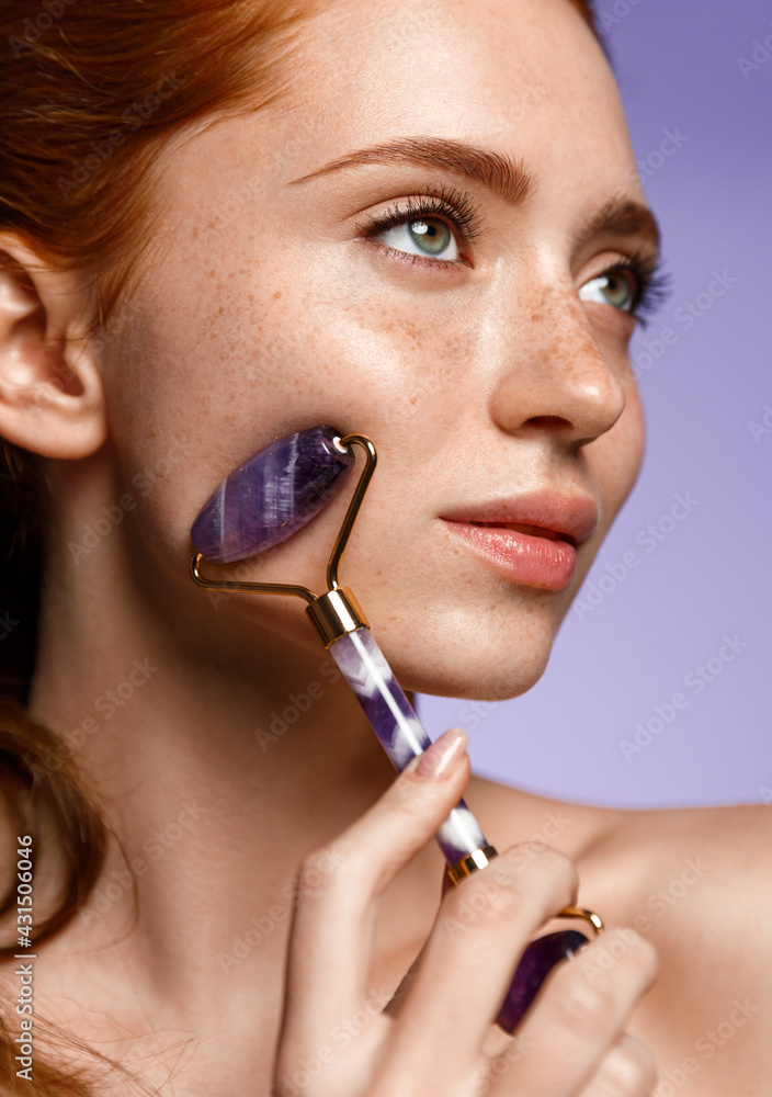 Close up of beauty redhead use natural jade face roller on cheek, massaging facial muscles, lifting, healthy glowing result. Girl doing spa skin care procedures, penetrates cream with quarz tool - obrazy, fototapety, plakaty 