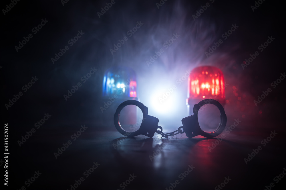 Police raid at night and you are under arrest concept. Silhouette of handcuffs with police car on backside. Image with the flashing red and blue police lights at foggy background.