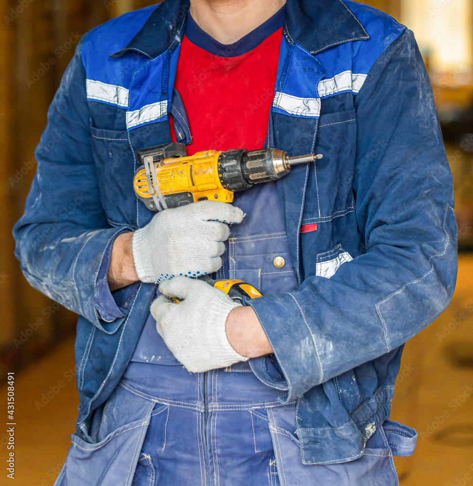 Fototapeta premium Worker in dirty uniform and protective gloves with drill in his hand in the room of apartment that is under construction, remodeling, renovation, extension, overhaul, restoration and reconstruction.