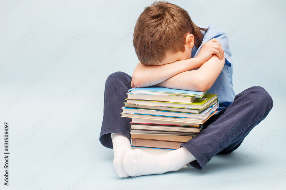Upset schoolboy sitting with pile of school books and covers his face with hands. boy sleeping on a stack of textbooks Upset schoolboy sitting with pile of school books and covers his face with hands. boy sleeping on a stack of textbooks