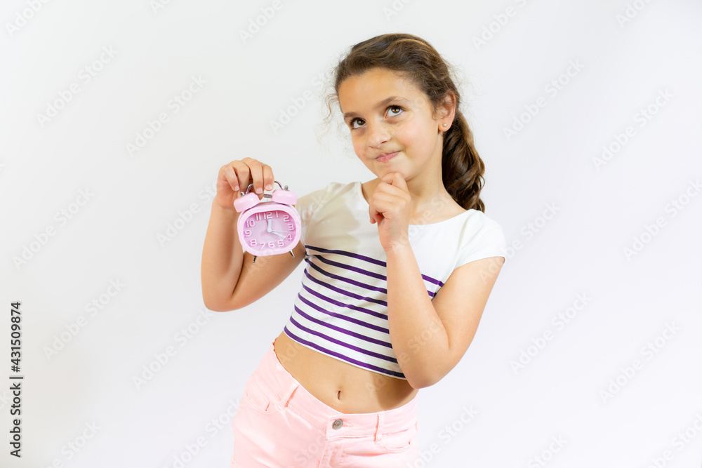 Thoughtful little girl brown-haired child touch chin with finger thinking or considering holding an alarm clock isolated over white background. Thoughtful little girl brown-haired child touch chin with finger thinking or considering holding an alarm clock isolated over white background.