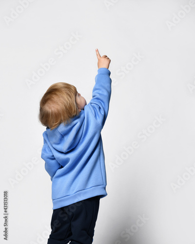 baby in a warm blue hoodie stands with his back to us and points something with his finger at the top
