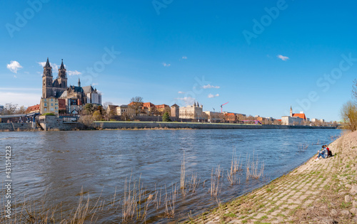 Panoramic view over Magdeburg historical downtown, Elbe river and the cathedral in early Spring with two girls sitting at the bank with warm illumination and blue sky, Germany.