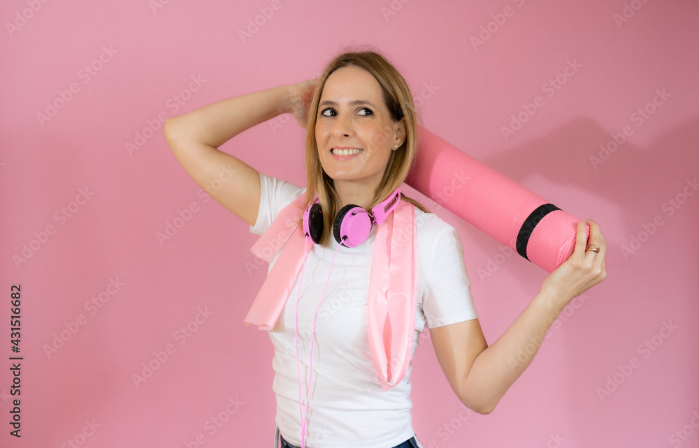 young woman holding a pilates mat and wearing headphone with a smile - isolated on pink young woman holding a pilates mat and wearing headphone with a smile - isolated on pink