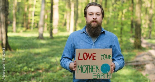 Portrait shot of Caucasian young male eco acivist standing in green forest and holding poster with words Love our Mother Earth. Handsome man protesting for clean and safe environment. photo