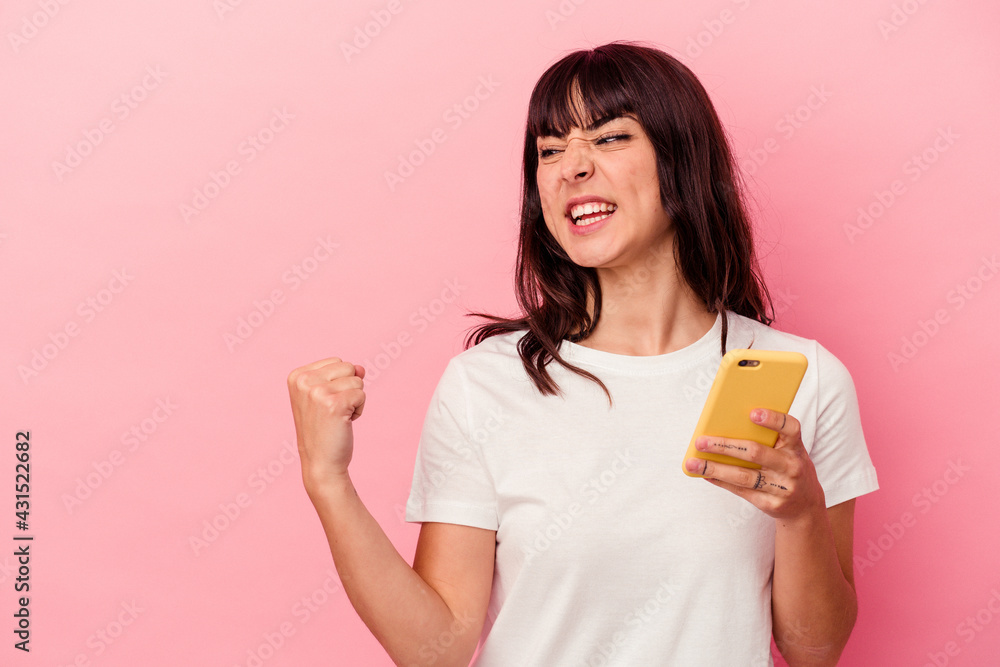 Young caucasian woman holding a mobile phone isolated on pink background raising fist after a victory, winner concept. Young caucasian woman holding a mobile phone isolated on pink background raising fist after a victory, winner concept.