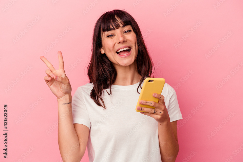 Young caucasian woman holding a mobile phone isolated on pink background joyful and carefree showing a peace symbol with fingers. Young caucasian woman holding a mobile phone isolated on pink background joyful and carefree showing a peace symbol with fingers.