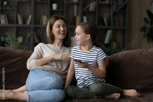 Overjoyed young Caucasian mom and small teenage daughter relax on couch at home have fun using cellphone together. Smiling mother and little teen girl child laugh browsing modern smartphone.