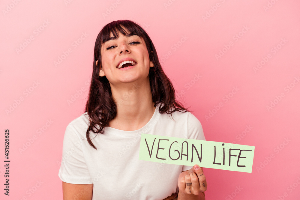 Young caucasian woman holding a vegan life placard isolated on pink background laughing and having fun. Young caucasian woman holding a vegan life placard isolated on pink background laughing and having fun.