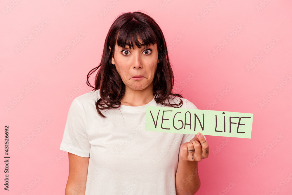 Young caucasian woman holding a vegan life placard isolated on pink background shrugs shoulders and open eyes confused. Young caucasian woman holding a vegan life placard isolated on pink background shrugs shoulders and open eyes confused.