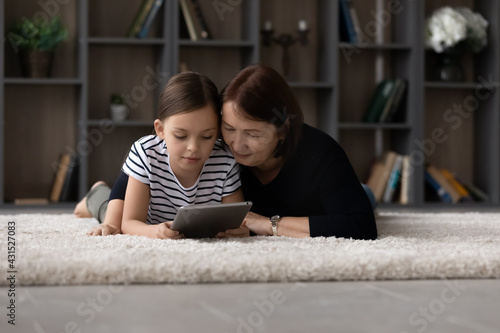 Loving mature grandmother and teen granddaughter relax at home using modern pad device together. Happy elderly granny and little girl child have fun browsing talking on video virtual call on tablet.