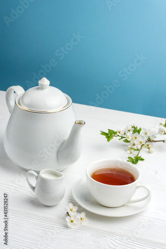 Teapot, tea cup, milk jug and cherry branch on a blue background with copy space.