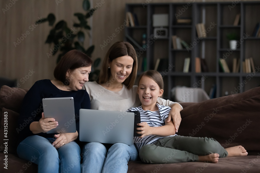 Happy modern three generations of Caucasian women sit relax on sofa at home using gadgets together. Smiling girl child with mother and granny have fun browsing devices. Technology concept. Happy modern three generations of Caucasian women sit relax on sofa at home using gadgets together. Smiling girl child with mother and granny have fun browsing devices. Technology concept.